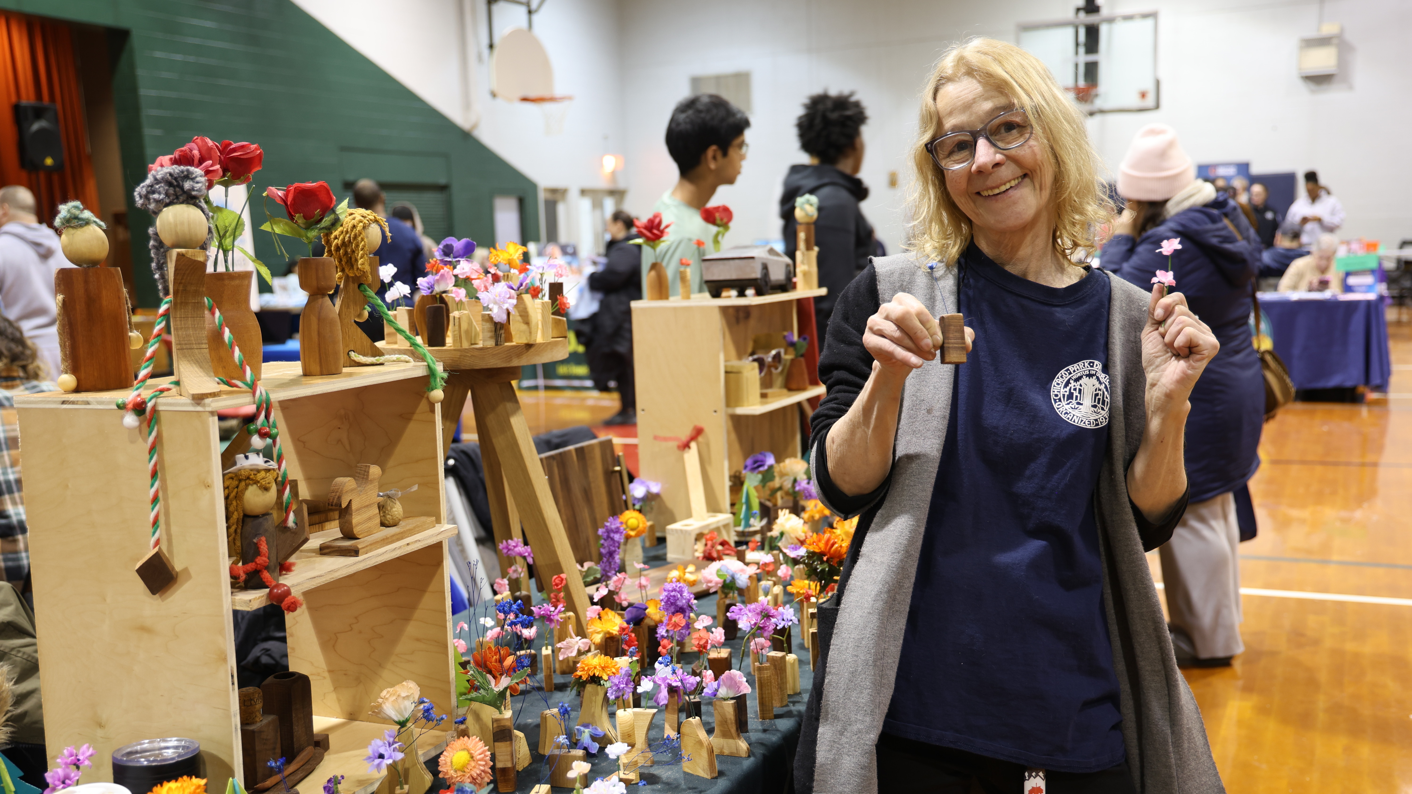 A smiling woman holds wooden crafts next to a table filled with tiny colorful floral arrangements.
